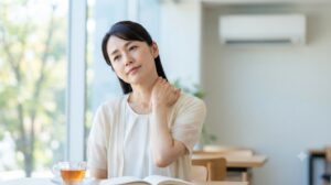 冷房の効いた室内で首や肩の重だるさを感じる女性。夏の寒暖差による首こり・肩こりをイメージした写真